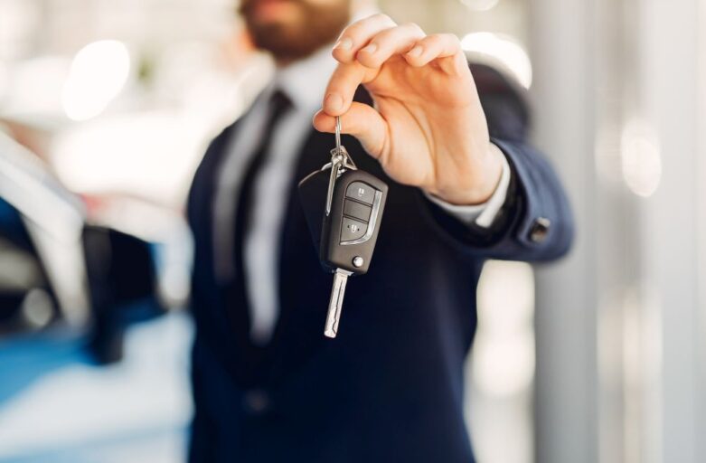 A person in a suit holds out a car key fob and key, with a blurred car visible in the background—perfect for showcasing seamless Singapore car rental services.