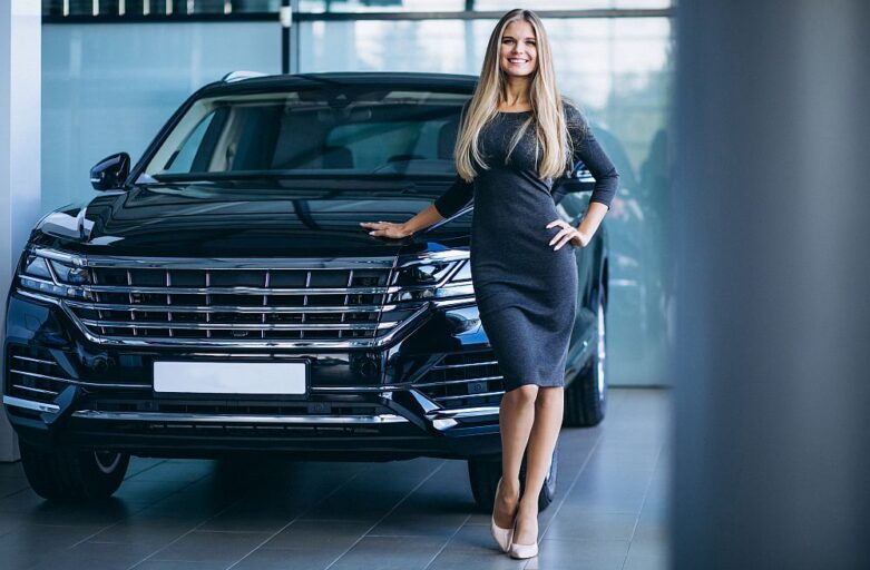 A woman in a black dress stands next to a black SUV inside a showroom, smiling at the camera—showcasing an exceptional luxury car rental experience.