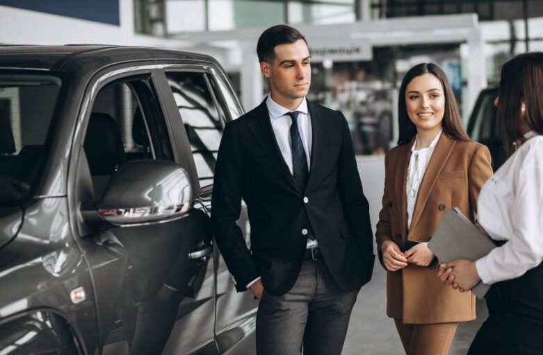 Two professionally dressed people stand next to a car in a showroom, discussing car rental in Singapore with a representative from singaporecarrental.sg holding a clipboard.