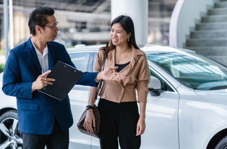 A man holding a clipboard gestures while speaking to a woman standing next to a white car inside a building, discussing options for the cheapest car lease Singapore has to offer.