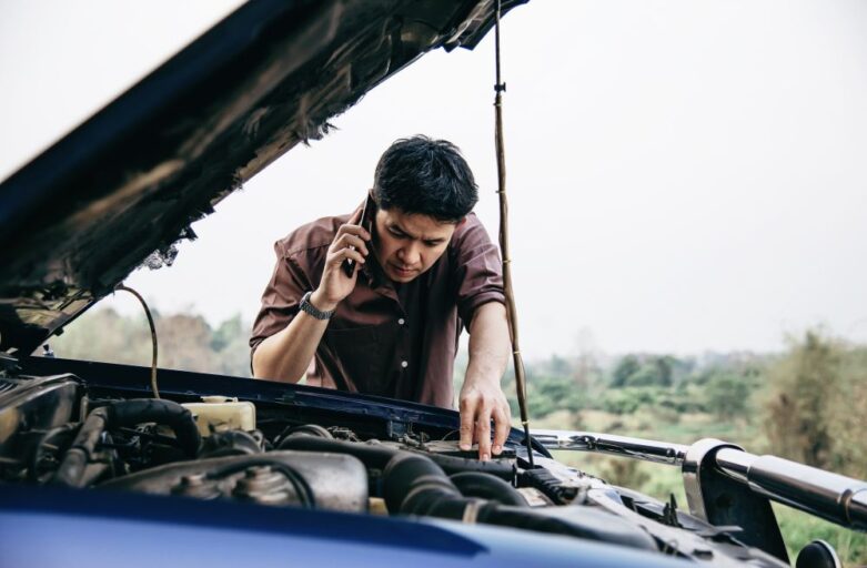 A man stands by a car with the hood open, talking on his phone while inspecting the engine outdoors, possibly discussing car transmission repair.