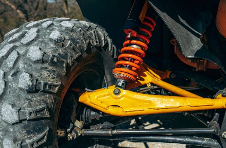 Close-up of an off-road vehicle's suspension and shock absorber system, featuring a large tire, orange coil spring, yellow control arm, and robust shock absorber.