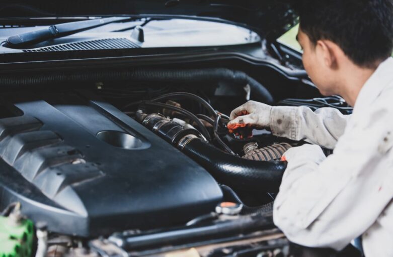 A mechanic wearing a white uniform works under the open hood of a car, inspecting the engine components.