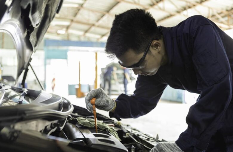 A mechanic wearing gloves and safety glasses checks the oil level under the hood of a car in an indoor auto repair shop.