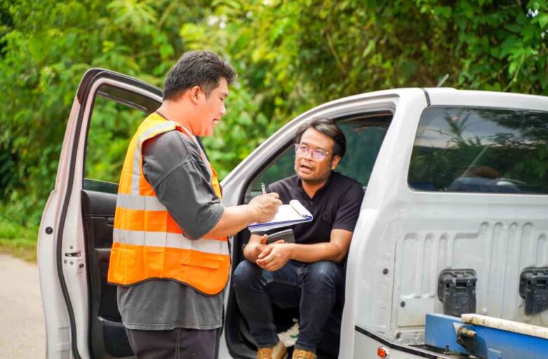 A man in an orange safety vest writes on a clipboard while another man sits on the open tailgate of a white pickup truck, surrounded by greenery.