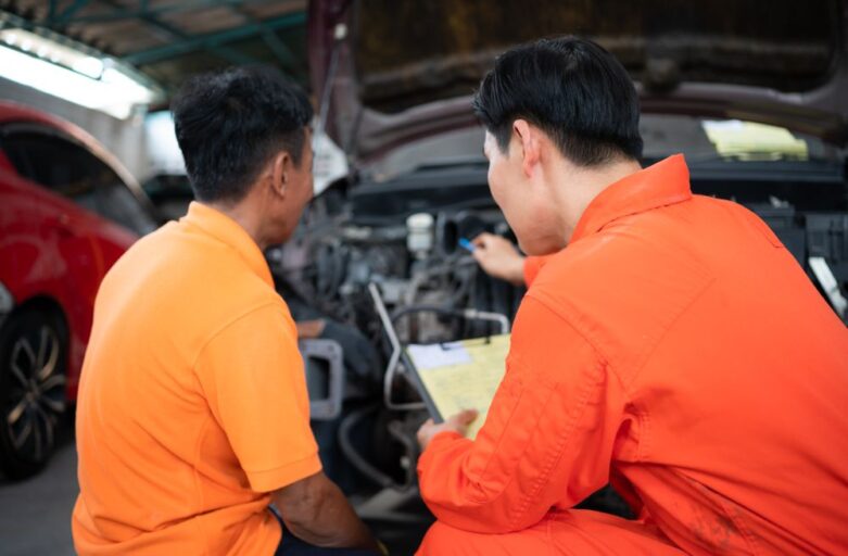 Two men examine a car engine in a garage; one holds a clipboard and points at the engine while discussing with the other, illustrating A Simple Guide to Common Car Repair Services.