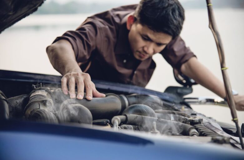 A man inspects the engine of a car with the hood open and visible smoke rising from the engine components—clear signs your car may need engine repair.